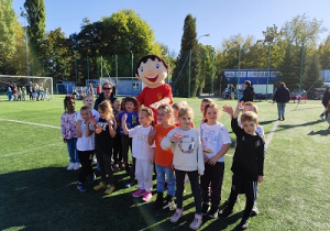 Dzieci bawią się z maskotką imprezy sportowej.