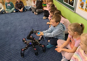 Dzieci siedzą na podłodze i podziwiają małego robota.