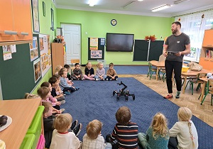 Dzieci siedzą w kole, w środku koła robot, prowadzący opowiada dzieciom o budowie robota